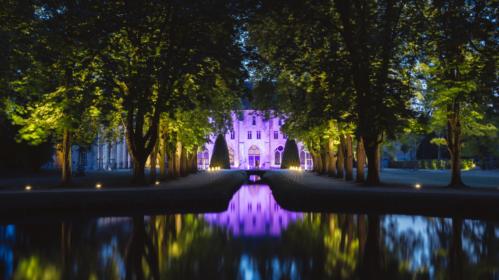 Photo du parc de Royaumont de nuit avec jeux de lumières sur la façade du monument et reflet de la vue dans l'eau.