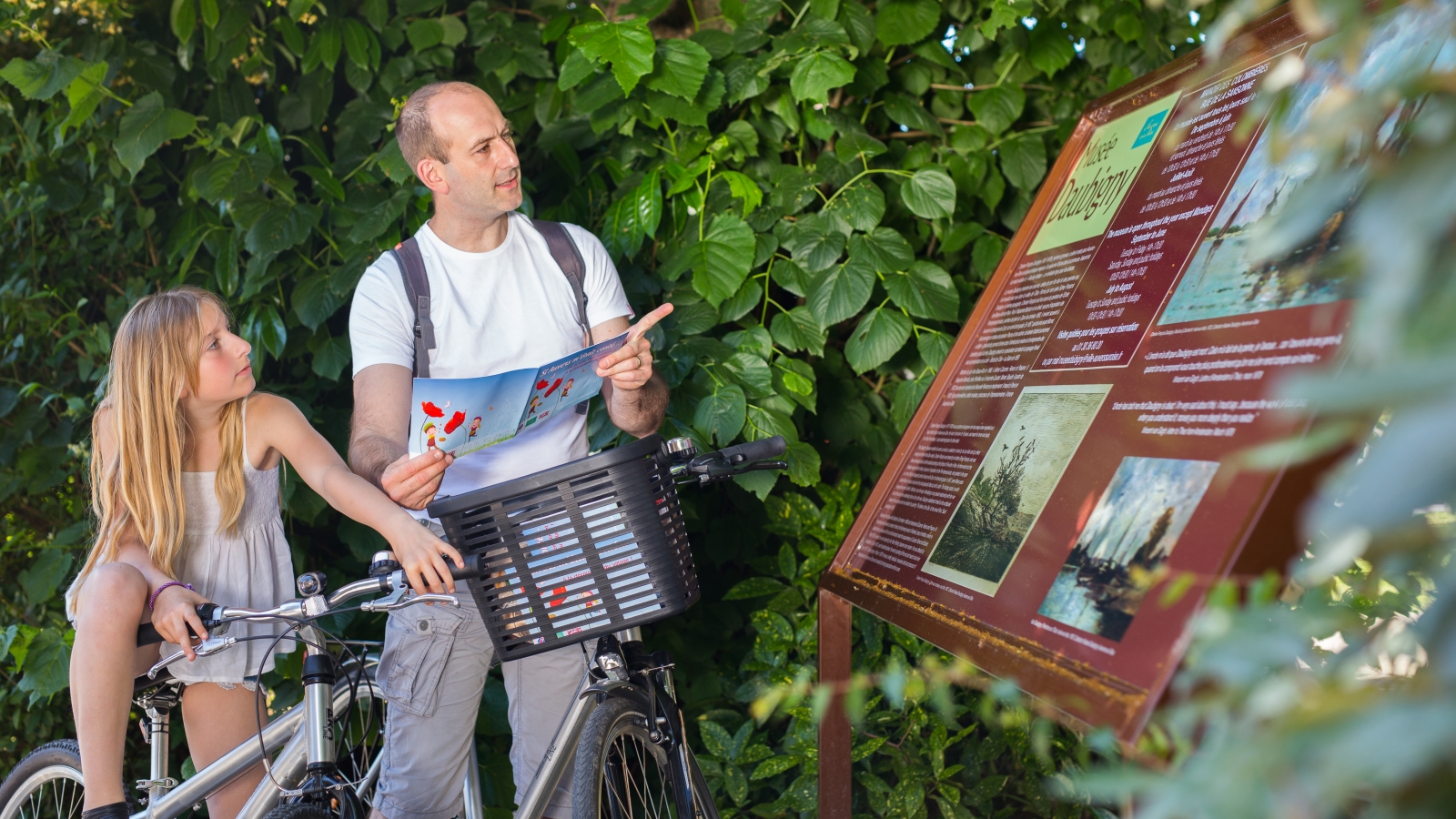 Photo d'un père et sa fille à vélo se renseignant sur un panneau du musée de Daubigny à Auvers-sur-Oise