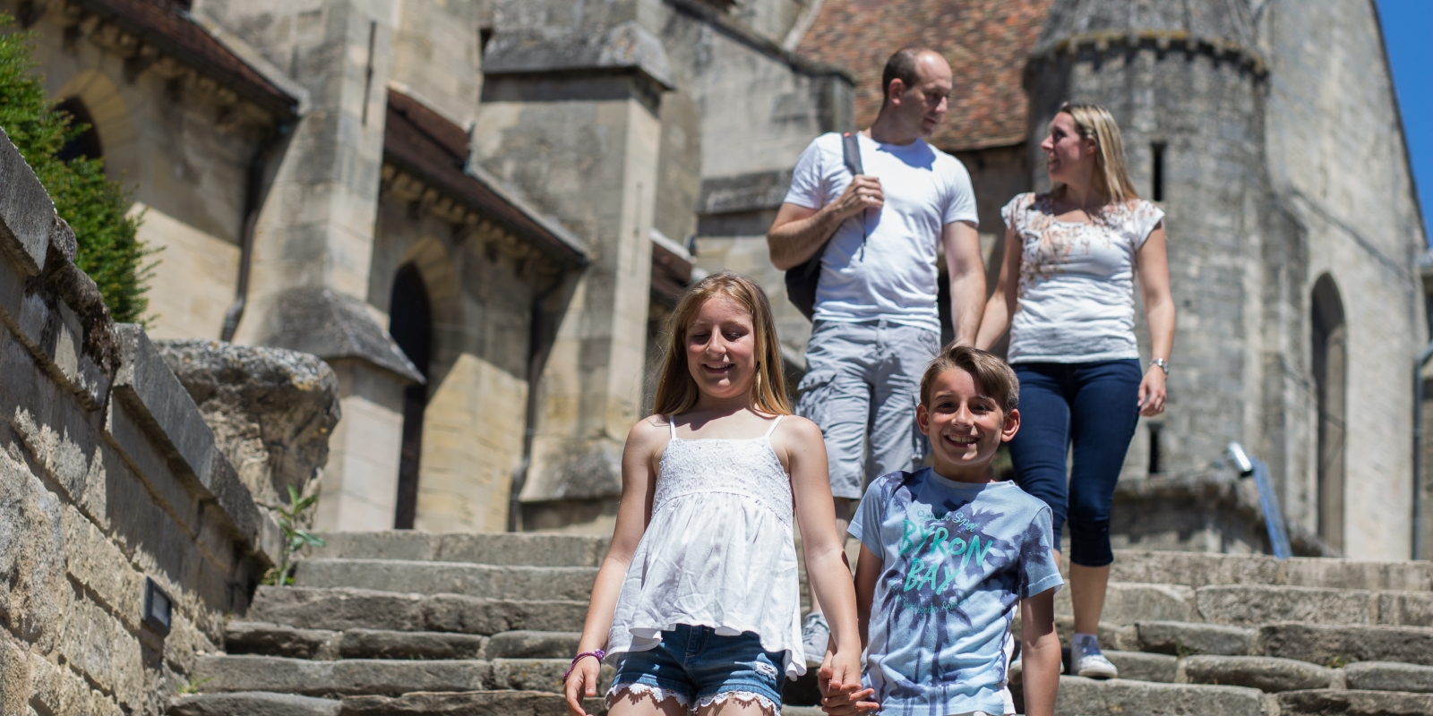 Photo d'une famille souriante avec deux enfants et leurs parents se tenant la main en descendant les marches près de la célèbre église d'Auvers sur Oise peinte par Vincent Van Gogh dans le Val d'Oise sous un beau ciel bleu