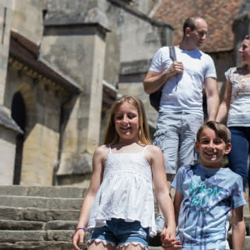 Photo d'une famille souriante avec deux enfants et leurs parents se tenant la main en descendant les marches près de la célèbre église d'Auvers sur Oise peinte par Vincent Van Gogh dans le Val d'Oise sous un beau ciel bleu