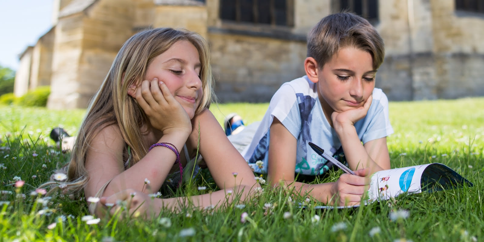 Photo de deux enfants dessinant sur un carnet de visite allongés dans l'herbe devant l'église d'Auvers sur Oise dans le Val d'Oise