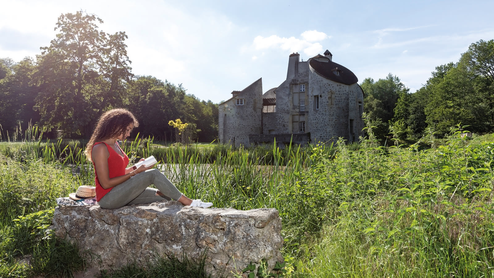 jeune femme avec un livre assise sur un muret devant le Château de la Chasse en arrière plan